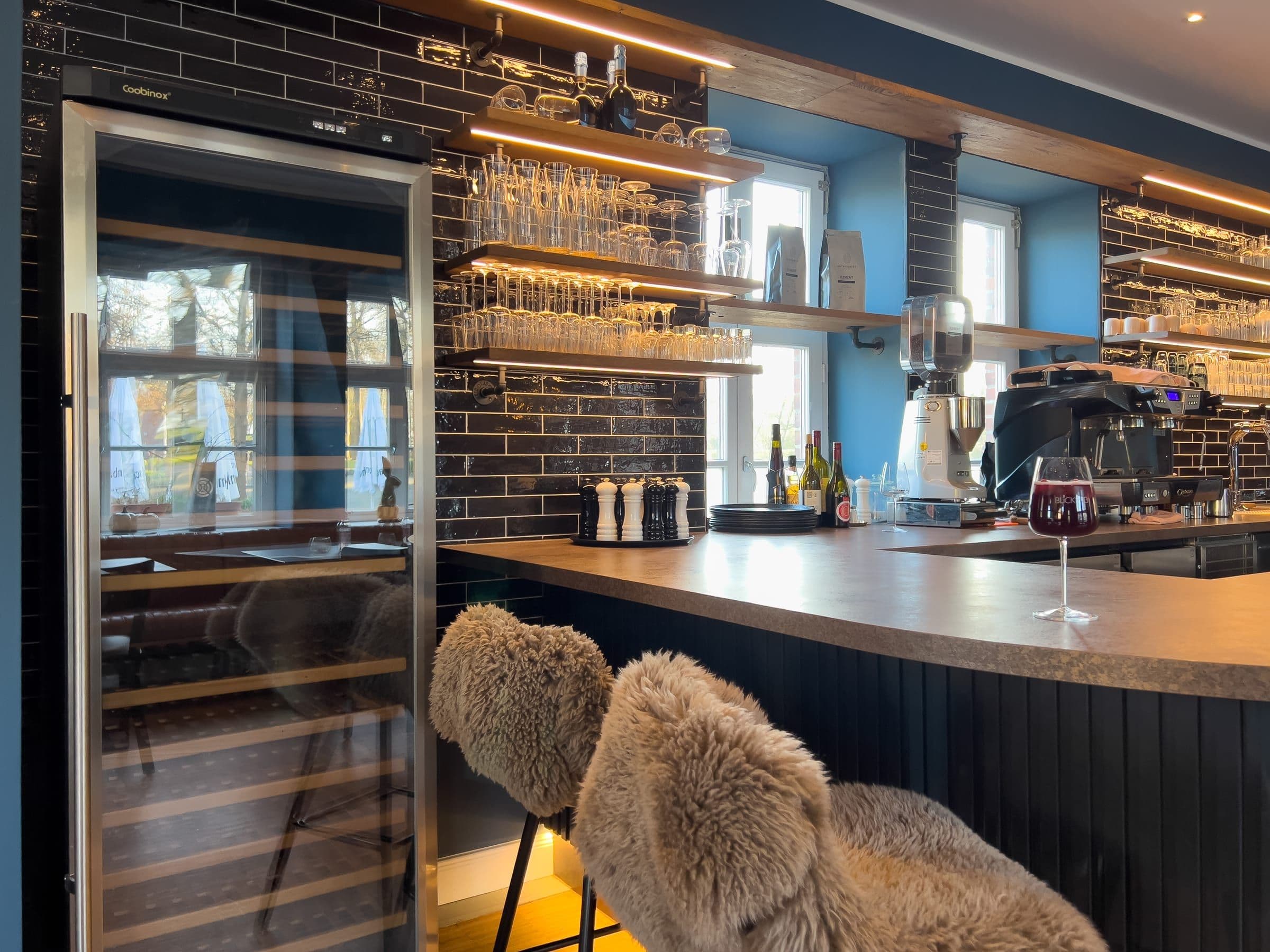 Bar — Bar im historischen Torhaus auf der Carlshöhe: dunkle Metro-Fliesen, Weinkühlschrank, Espressomaschine und Stehhocker mit Fellauflage