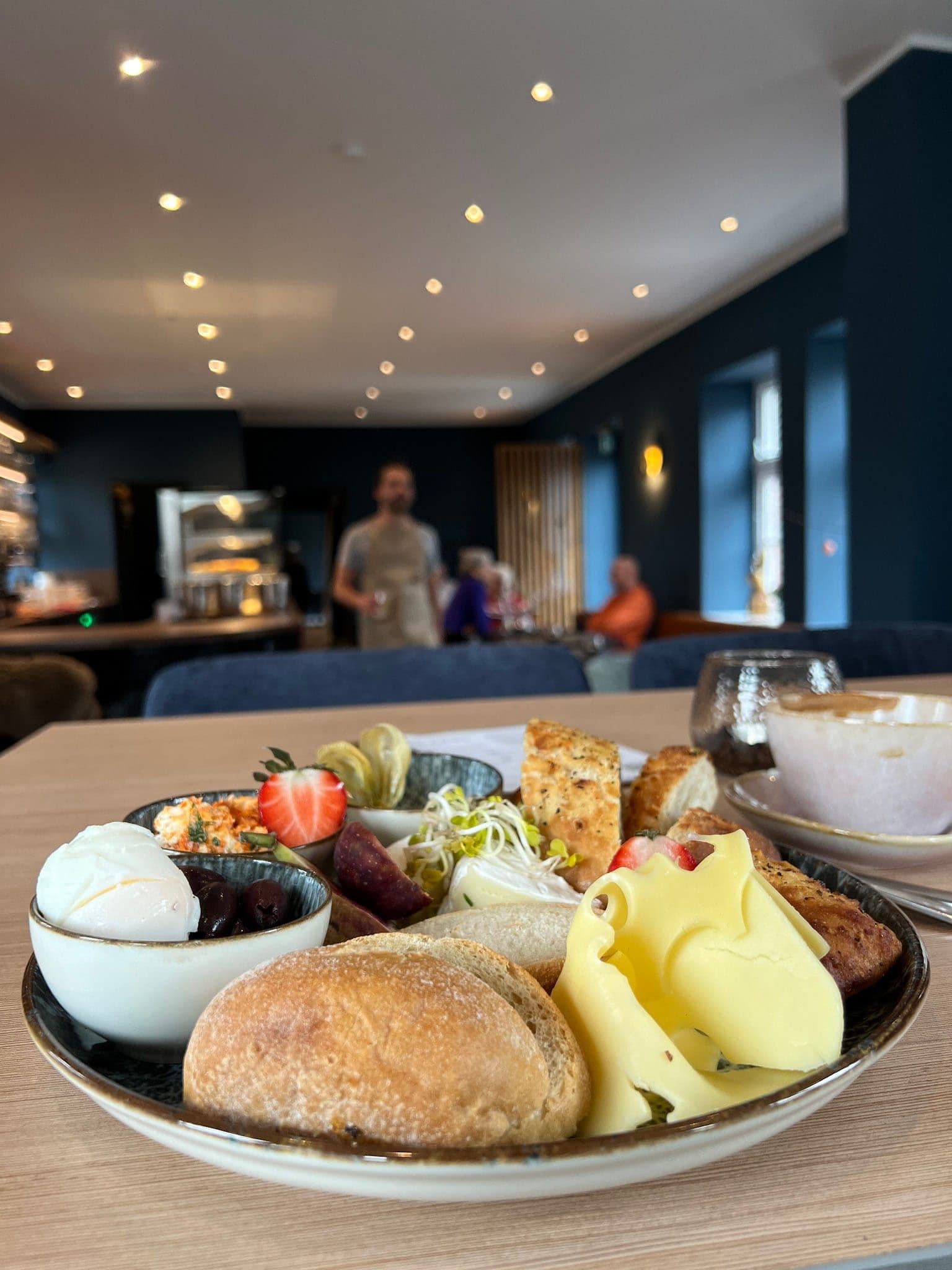 Frühstücks-Teller im BLICKFANG: Brötchen, drei Käsesorten, Aufschnitt, weiches Ei mit Oliven, Erdbeeren, marinierter Feta — formatfüllendes Tellerportrait